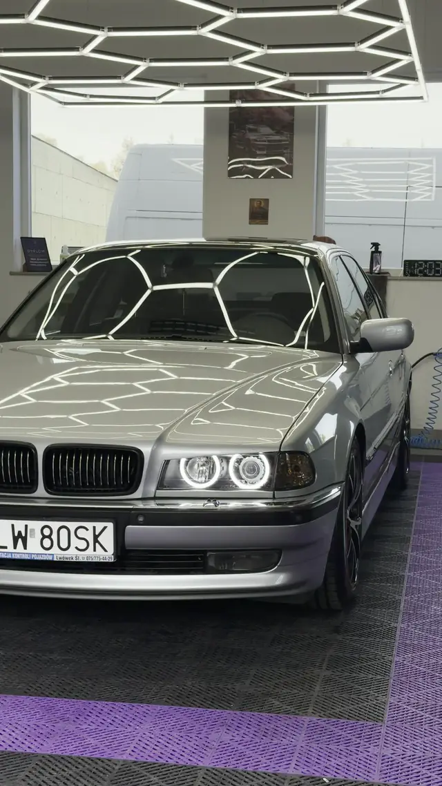Srebrne BMW serii 7 E38 w hali, na masce odbijają się sześciokątne lampy sufitowe