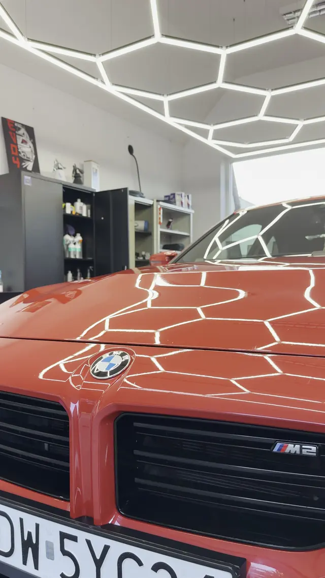 Czerwona maska BMW M2 w hali, na lakierze odbija się sześciokątna lampa sufitowa